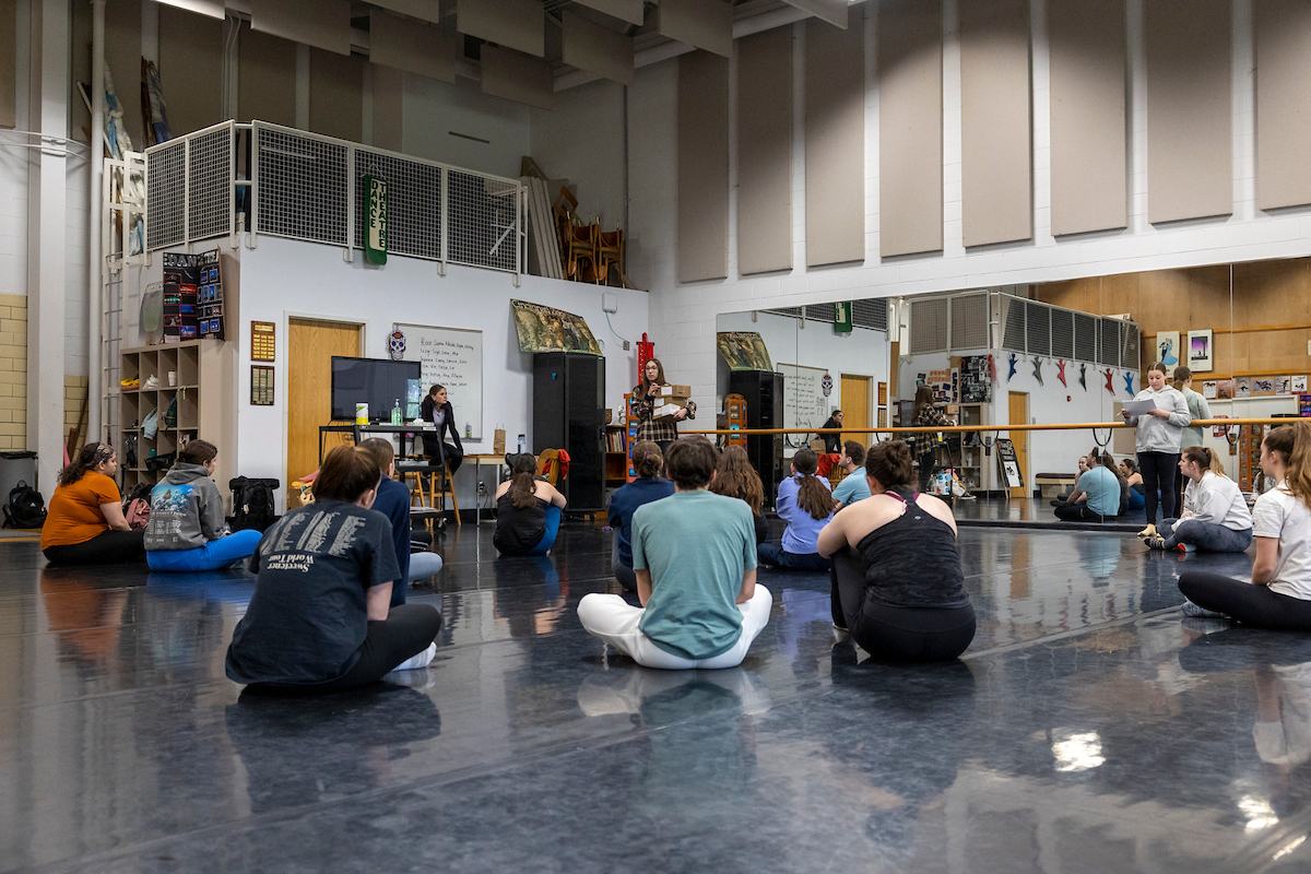 Students sit on the floor of a dance studio listening to a professor.