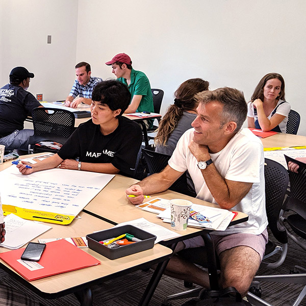 Faculty and graduate students sit at tables in groups of four. They work together on an activity and write on large post-it boards.