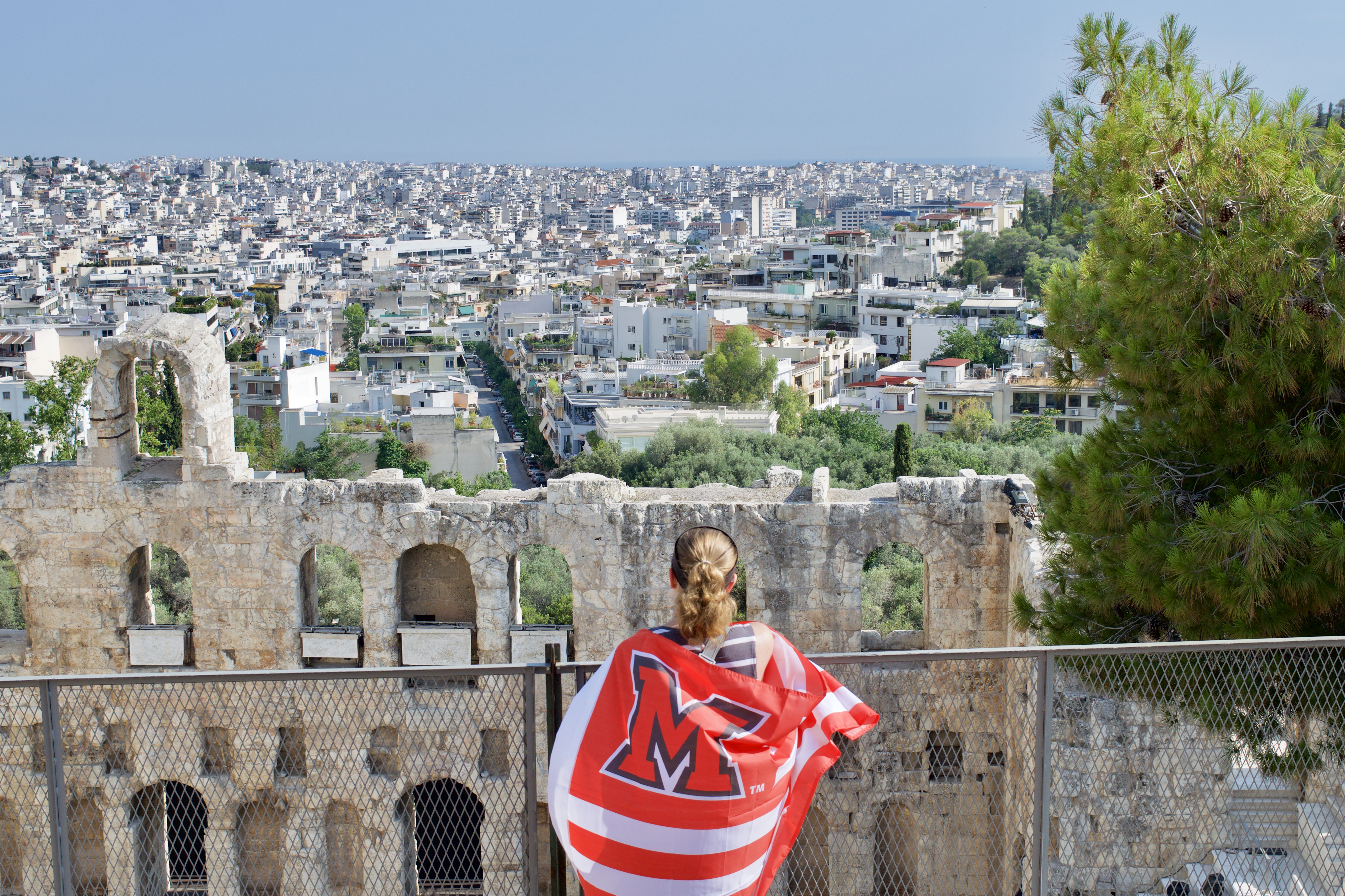A person stands overlooking Athens, Greece, with a red Miami flag draped over their shoulders