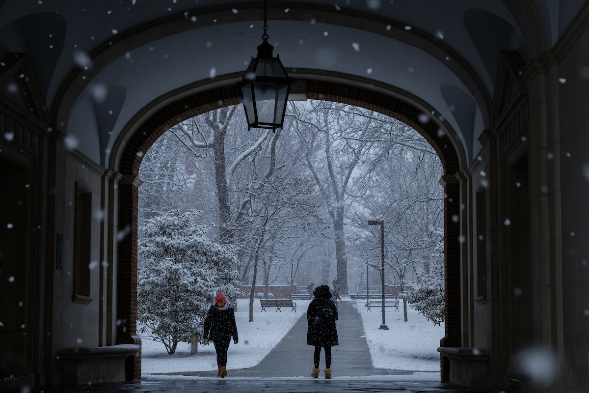 Close up of Upham arch, on the other side is two people walking, one towards the camera and one away. Behind them is a snowy scene and in front of the camera are snowflakes in the air