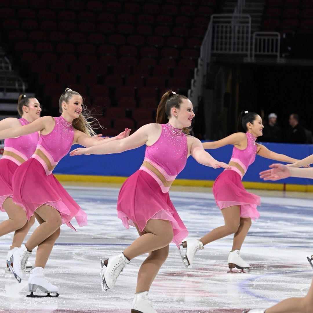 Shot of 4 figure skaters, all with tight ponytails and wearing sparkly pink uniforms with white skates. Xandra skates in the middle