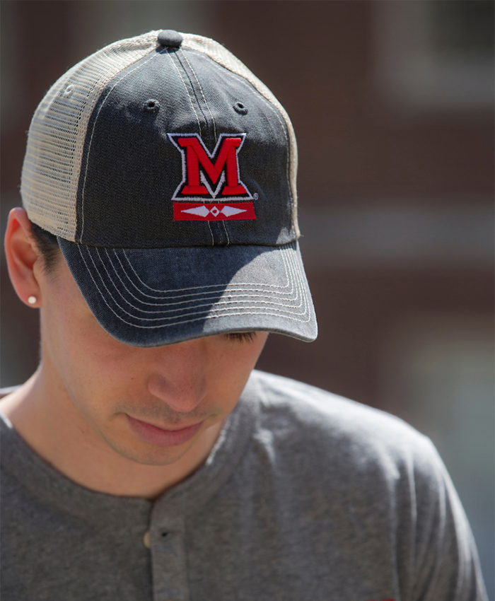 A student looking downward wears a grey and white trucker cap, with large red 'M' and Myaamia logo