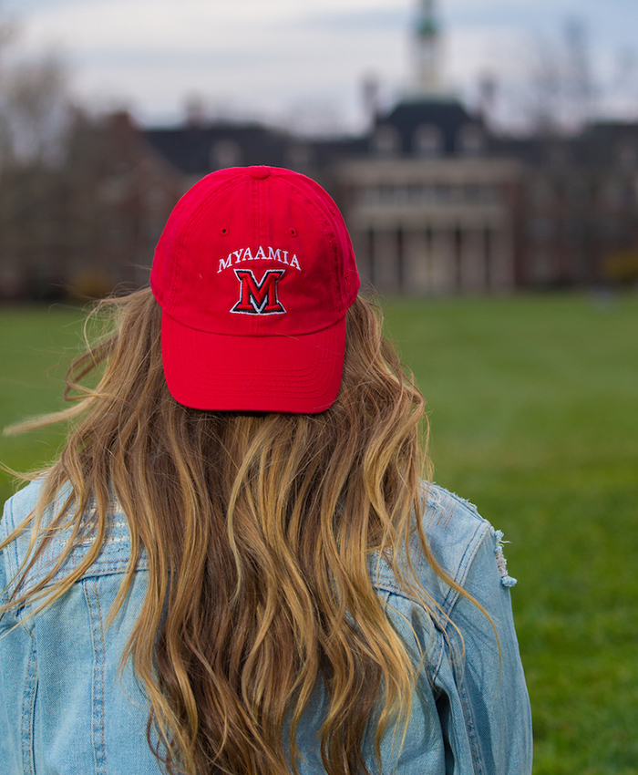 Student sits looking across Central Quad and wearing a red baseball hat. Design includes the Miami University block M logo and the word 'myaamia' above it.