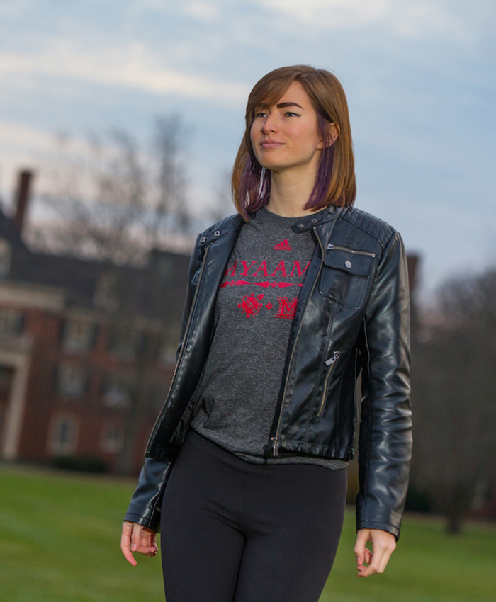 Student wearing a grey t-shirt under a black jacket. The shirt design includes the myaamia heritage logo, Miami University logo, the Adidas logo, and myaamia turtle mark. The word 'myaamia' is above.