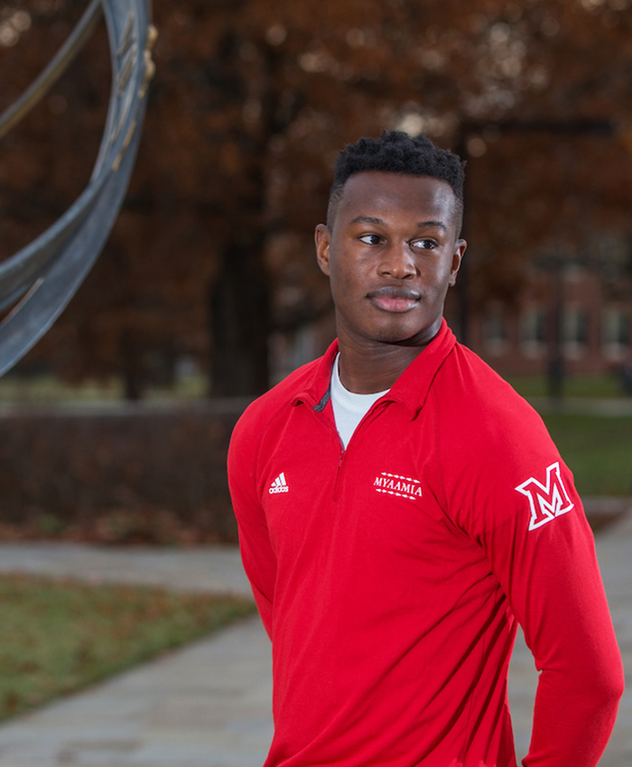  Student stands next to the sundial on central quad. He wears a red quarter zip pullover. The shirt design includes the myaamia heritage logo, Miami University logo, the Adidas logo, and the word 'myaamia' 