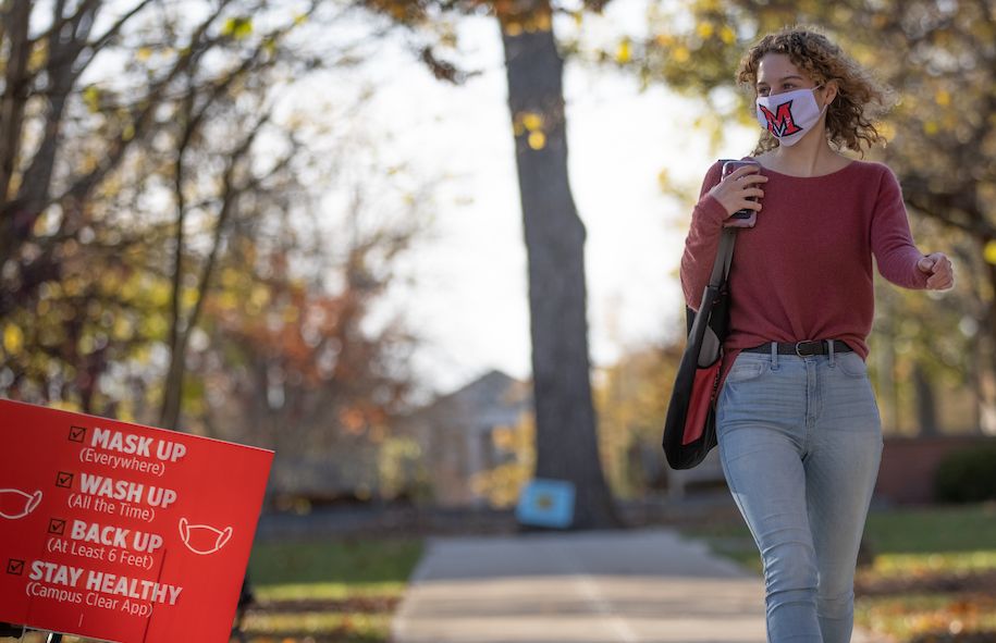 A student walks past a sign on campus that says Mask Up Everywhere, Wash Up all the time, Back Up at least 6 ft, Stay Healthy use the Campus Clear App