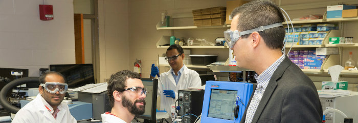 Professor Scott Hartley, wearing protective glasses, talks with students in the lab, CAS