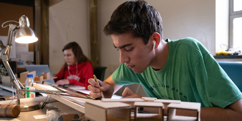 male student drawing at desk with model in front of him