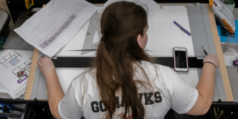  Viewed from behind, a female student is looking at her drawings on her desk