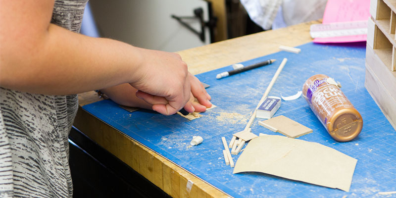  close-up view of hands cutting a piece for a model. On the worktable are scraps of material and a bottle of glue on its side