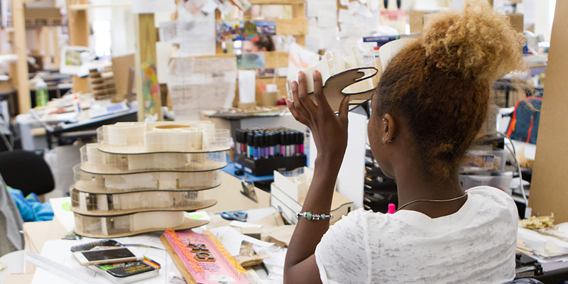 Viewed from behind the chair, a seated woman holds up a section of an architectural model and studies it intently, Other pieces and tools for the model are on the table in front of her.