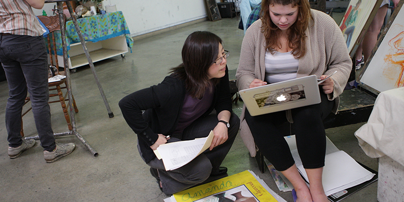 Joomi Chung consults with a student using a laptop as others paint at easels in the studio