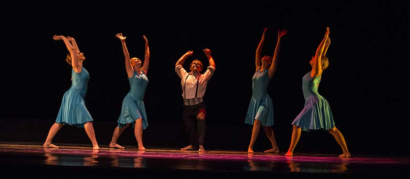 On a dimly lit stage, four female figures flank a male figure, all with arms raised