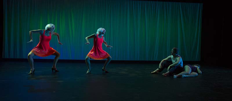 Two dancers in red dresses and blonde wigs move toward figures on the floor of the stage