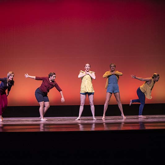 Six dancers perform. Some lean forward and others cross arms across their chests.