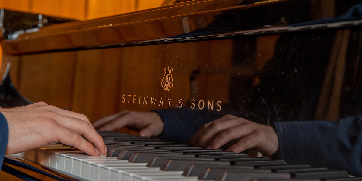 Hands on a keyboard are reflected against the mirrored surface of the Steinway grand piano