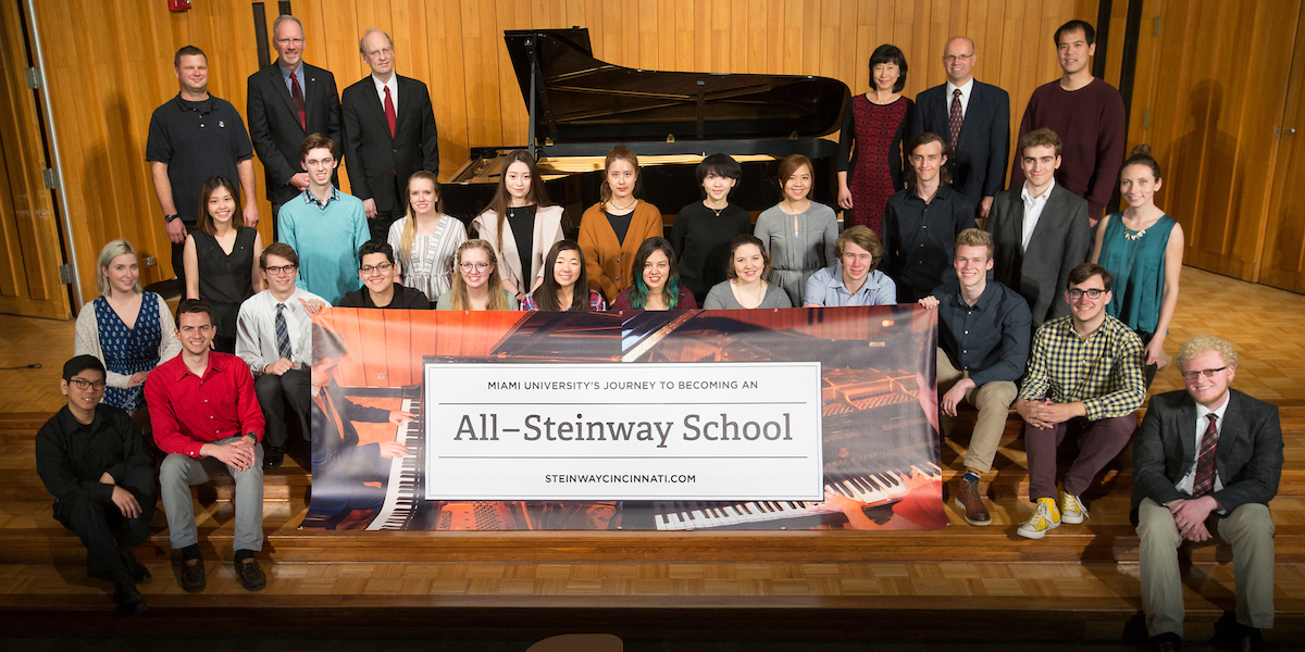 A group of students and faculty gather onstage in front of a Steinway Grand at Souers as they hold a banner announcing that Miami is in process of becoming a Steinway School