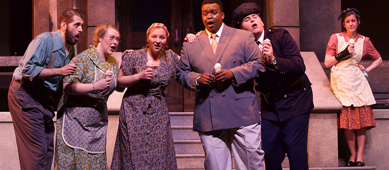 A group of singers holding ice cream cones in front of a brownstone building. A woman wearing an apron stand off to the side in apparent dejection in scene from the Miami Opera production of Street Scene Musical
