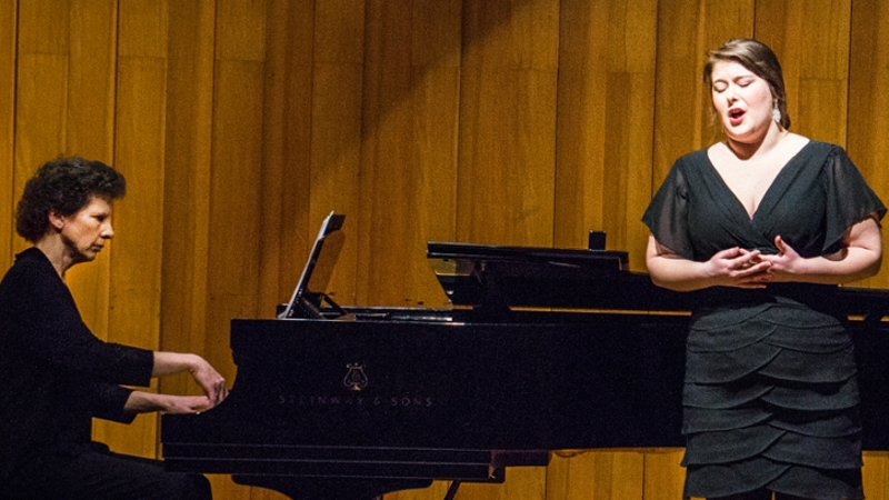 A woman closes her eyes and clasps her hands as she sings during a recital. Her accompanist and the grand piano are in the background