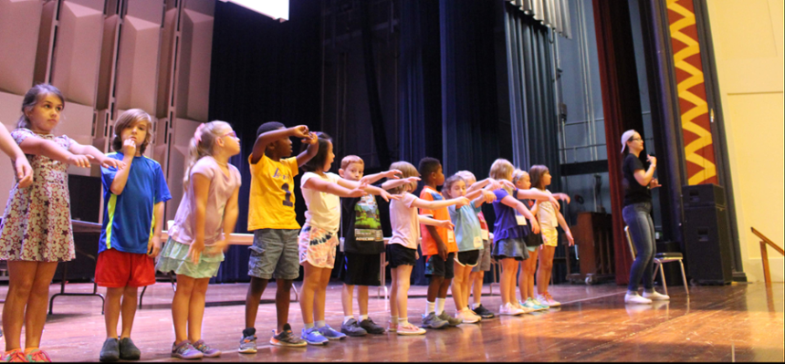 A row of children across the front of the stage, with an adult facilitator