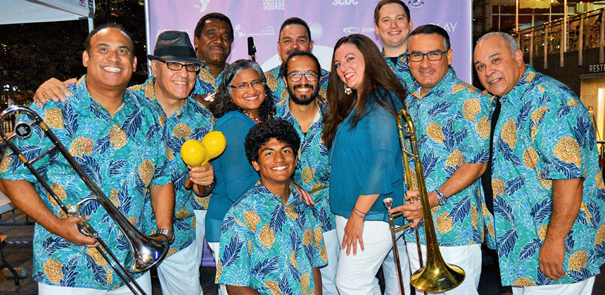 The 11 members of the Son Del Caribe Ensemble in colorful matching shirts of turquoise and yellow. Two are holding trombones 