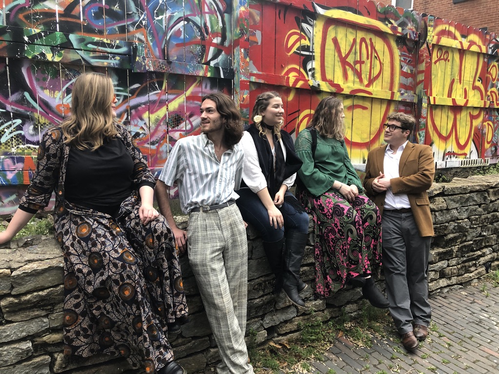 Actors three women and two men in sixties era cloths having a conversation in front of a graffiti covered wall