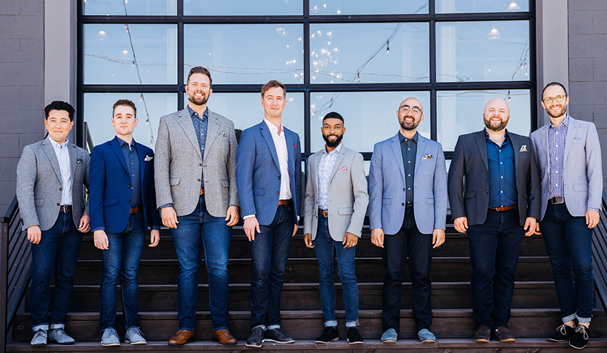 A professional group photo of the members of Cantus eight adult men wearing sport coats, button down shirts and dark jeans in colors of blue and grey