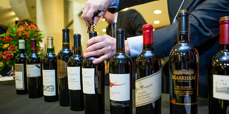 Hands are seen using a corkscrew to open a bottle, with other bottles of wine arrayed around it.