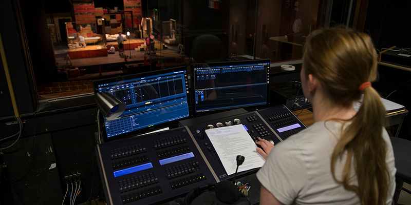 Looking toward the illuminated stage, a student operates a lighting board in Gates Abegglen Theatre