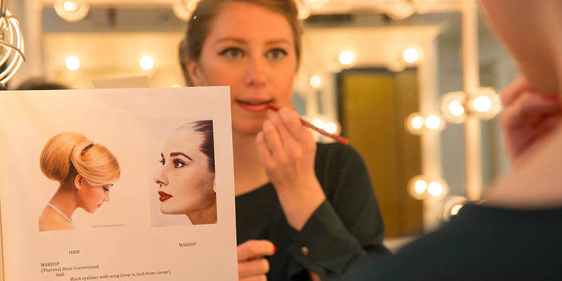 A student sits at a mirror and applies makeup by referring to a design