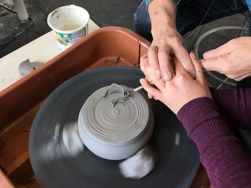 student's hands being guided by another to shape pottery on pottery wheel
