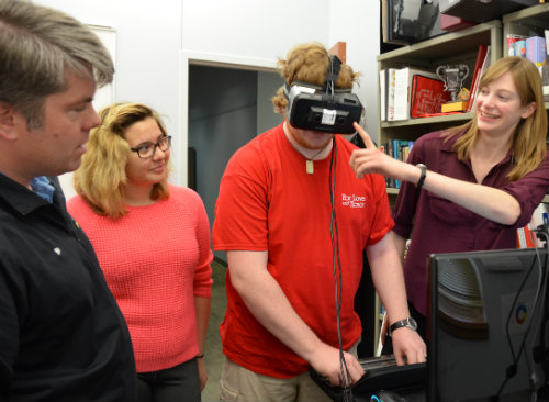 Two students and Dr. Brinkman watch as one student is wearing a virtual reality device and is preforming tasks