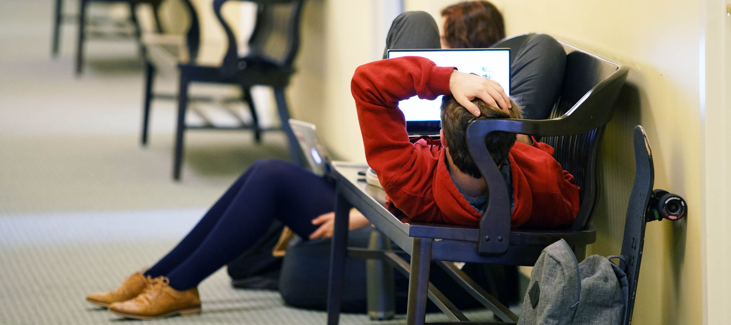 A student uses his laptop while reclined on a bench in a second-floor hallway