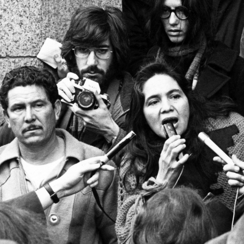Dolores movie scene; black and white; woman speaking into many microphones, news folks around her holding the microphones and cameras