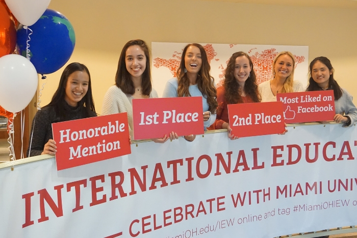 students holding signs that say "first place", "second place", etc. in MacMillan Hall's lobby, red and white balloons, banner on stairwell that says "International Education Week"