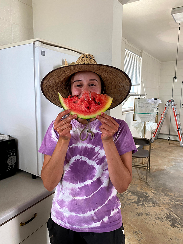 Grace stands in a kitchen, holding a curved slice of watermelon as a 'smile' in front of her face