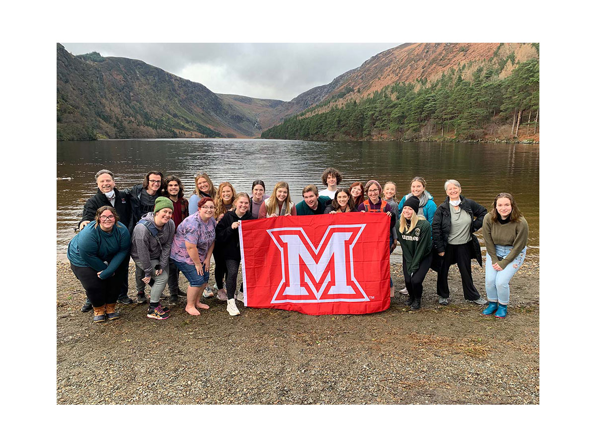 Students on a past Theatre in Dublin trip pose near a lake and hold up a Miami M flag