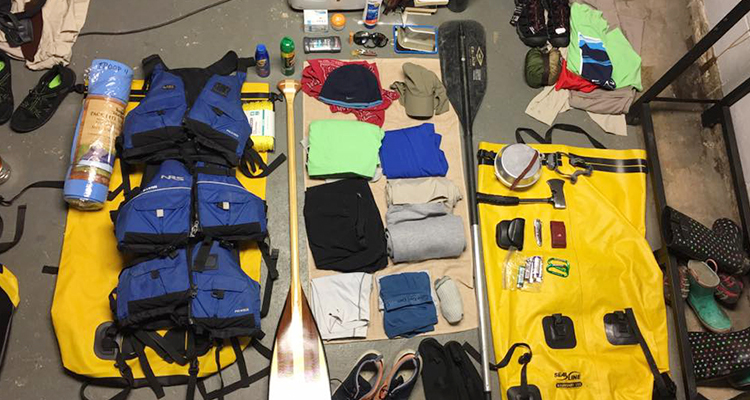 Canoeing gear spread out on the ground–life vests, clothes, a sleeping mat, sunblock, bug spray, paddles, an ax, sunglasses, watershoes, and more.