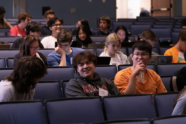 A student smiles while sitting with friends in a lecture hall.
