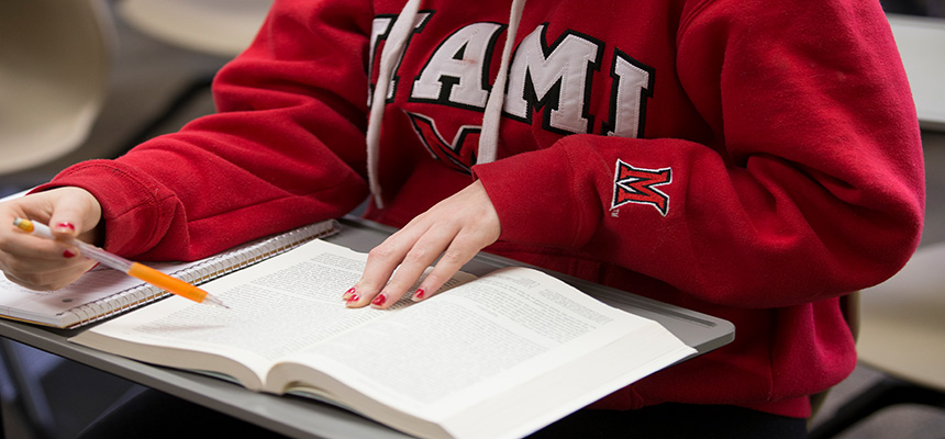 Image of a textbook on a desk with a pencil in someones hand and a Miami red sweatshirt