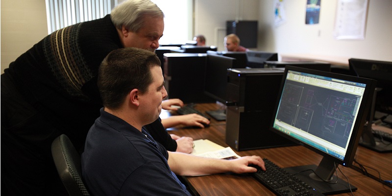 A student works at a computer in the computer lab while another man looks on