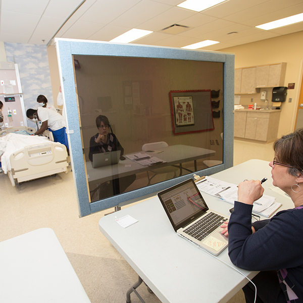 A professor watching a group of students work on the simulation manikin through a computer.