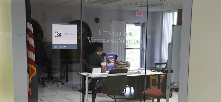  The glass doors of the veterans center with a student working at a desk inside.