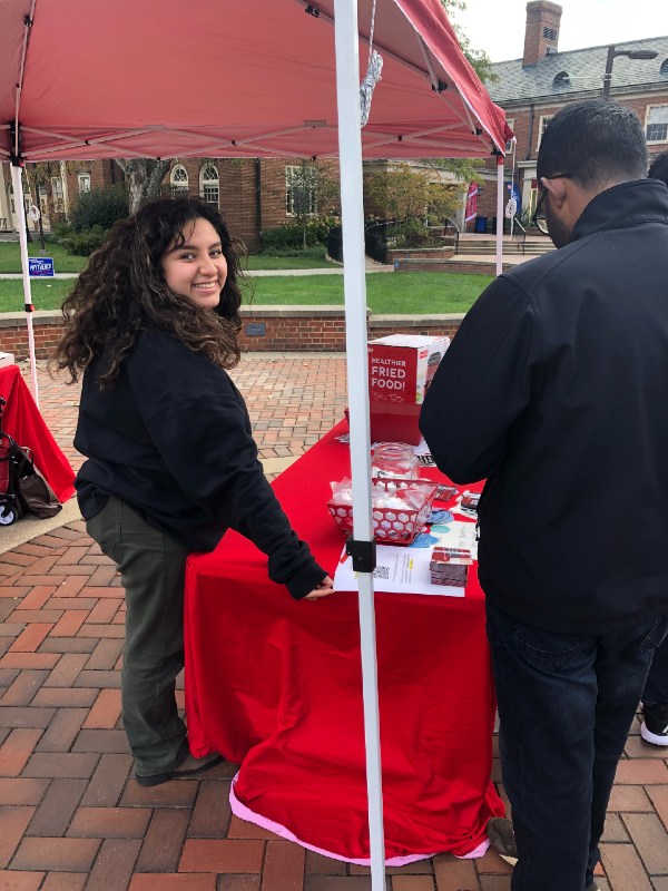 Student outside Shriver under a red tent with a table of information for Pizza at the Polls