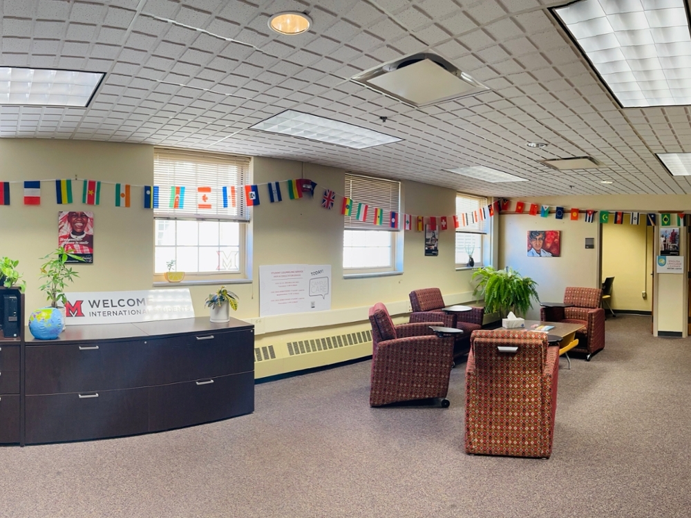 Room with armchairs and attached moveable trays, desks, and computers. The walls are lined with strings of international flags. A sign reads Welcome International students. 