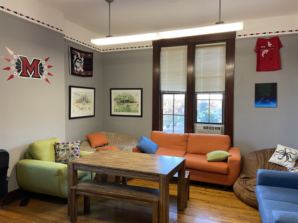 Colorful room with hardwood floors, comfortable couches, a bean bag chair, and wooden table and bench. Walls are decorated with illustrations, graphics, and t-shirts related to the Miami Tribe of Oklahoma. 