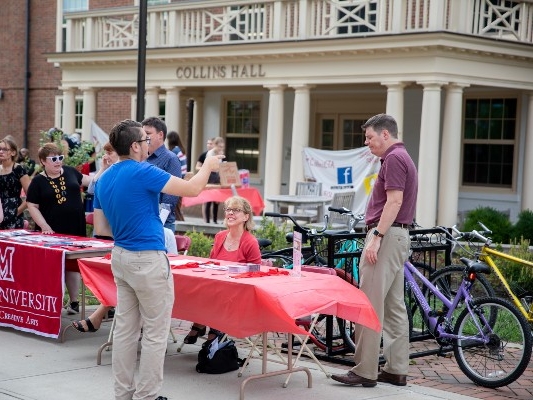 In front of Collins Hall, 7 people conversing around two information fair tables