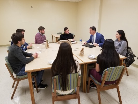 7 students, 1 faculty member seated and talking around a large square table