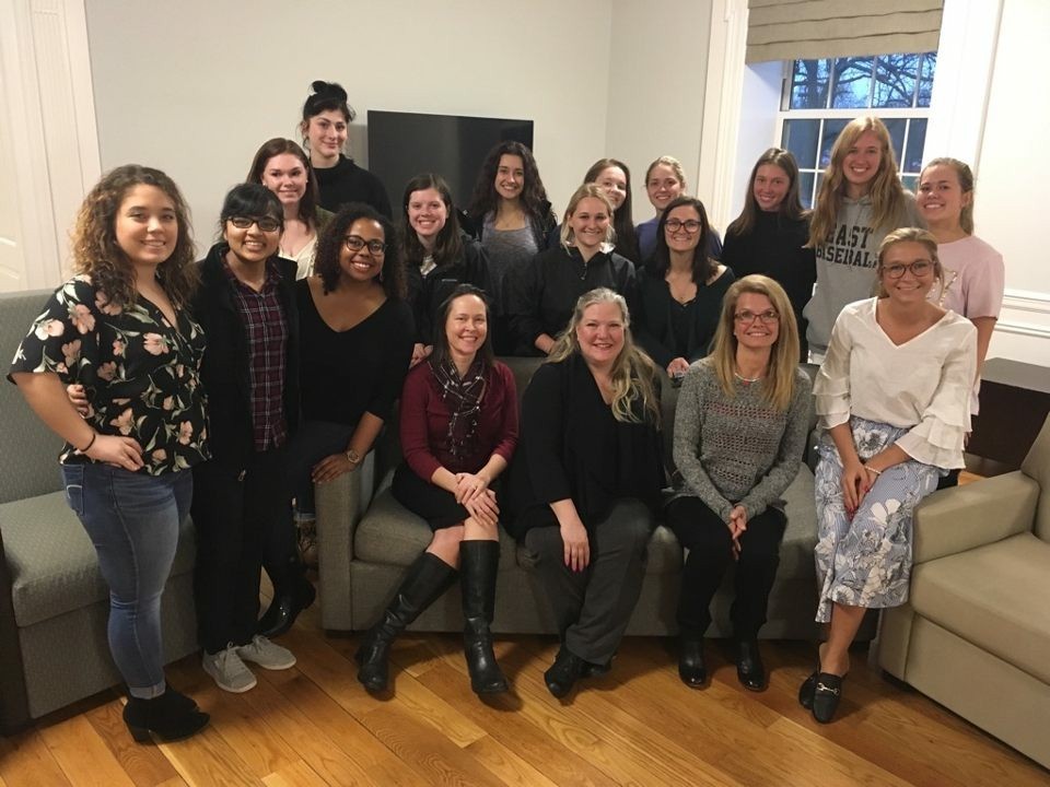 18 people posing for a picture in a lounge, some seating on a couch, some standing behind