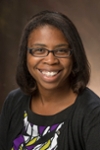 Vicka Bell-Robinson is an African American women with shoulder length hair who wears glasses. She is wearing a red top and standing against a brick colored background. 